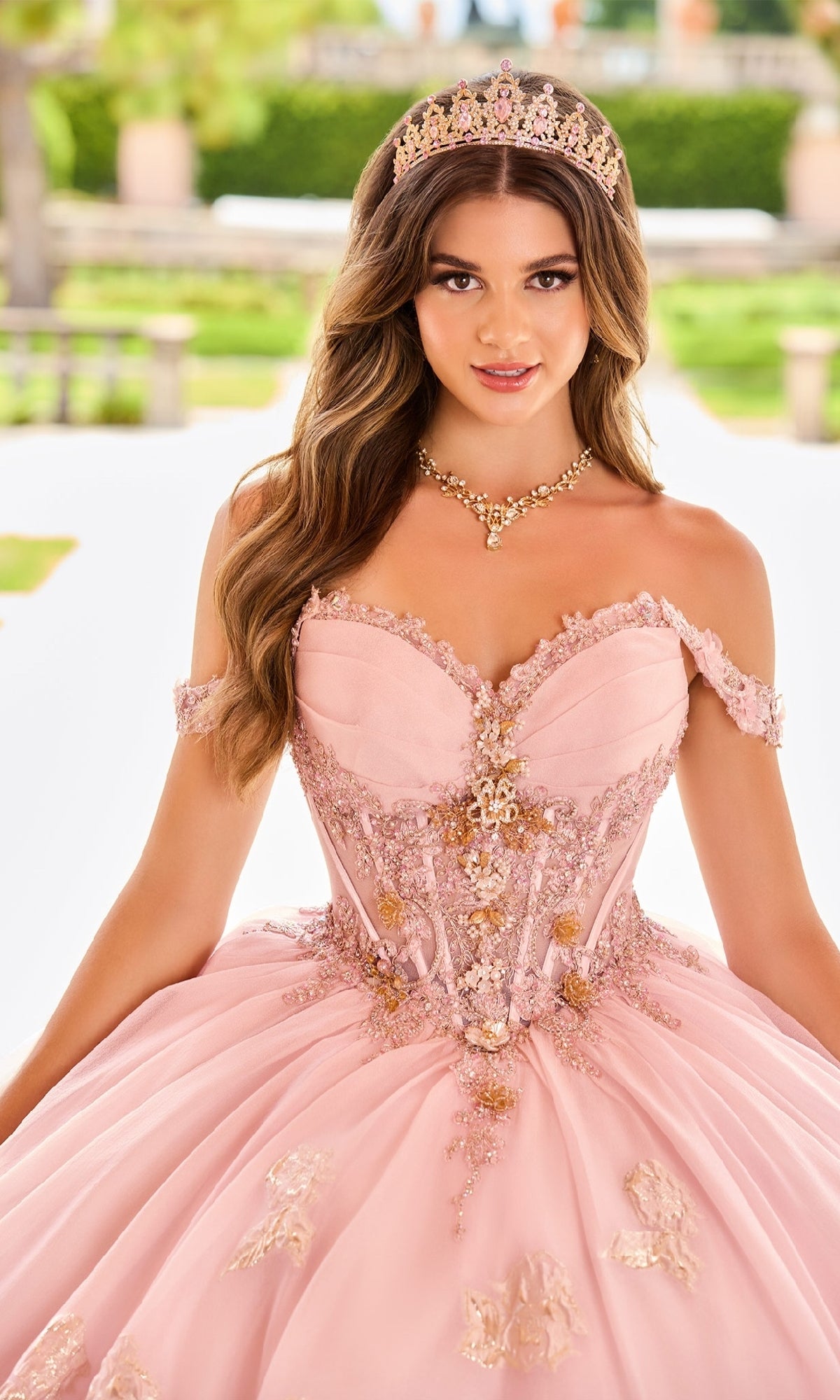 A young woman stands outdoors among lush greenery, wearing the Quinceanera Dress PR30154 By Princesa—a pink embellished ball gown with a sweetheart neckline—paired with a gold tiara.